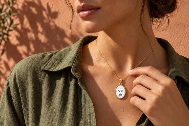 Modella mediterranea, capelli scuri raccolti in uno chignon morbido, sguardo basso, ciondolo personalizzato con frase incisa al collo su catenina sottile, luce naturale da finestra, sfondo lino crema
