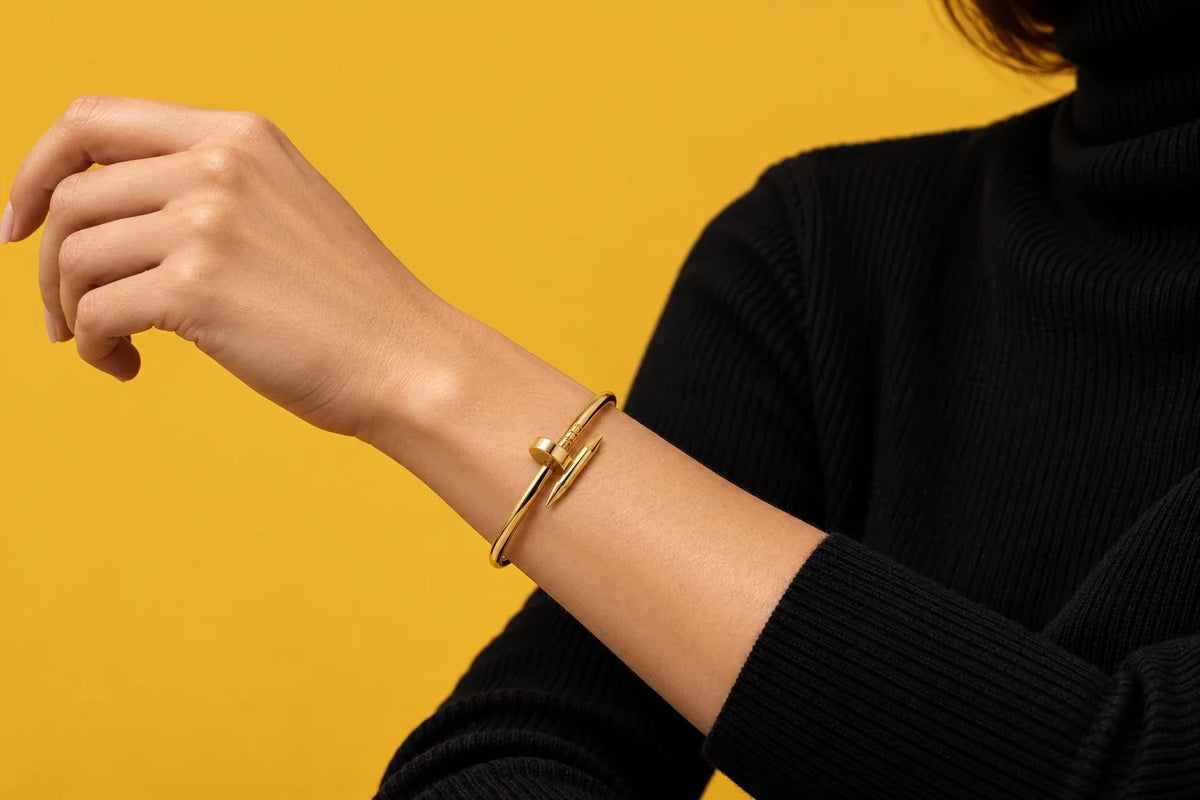 Modella mediterranea in maglione a trecce color avena, manica arrotolata, bracciale chiodo dorato sul polso sottile, luce invernale di finestra, sfondo intonaco caldo