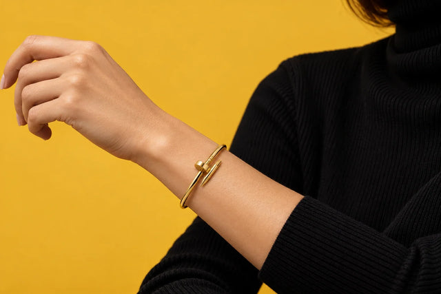 Modella mediterranea in maglione a trecce color avena, manica arrotolata, bracciale chiodo dorato sul polso sottile, luce invernale di finestra, sfondo intonaco caldo